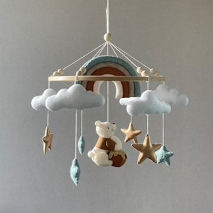 May include: A baby mobile featuring a rainbow, clouds, stars, and a bear holding a honey pot. The mobile is crafted from felt and suspended from a wooden ring.