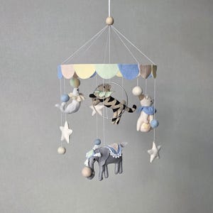 May include: A whimsical baby mobile featuring felt circus animals, including a tiger, elephant, seal, and bear. The mobile has a pastel-colored canopy and is suspended by strings with star and ball accents. The overall design is soft and inviting.