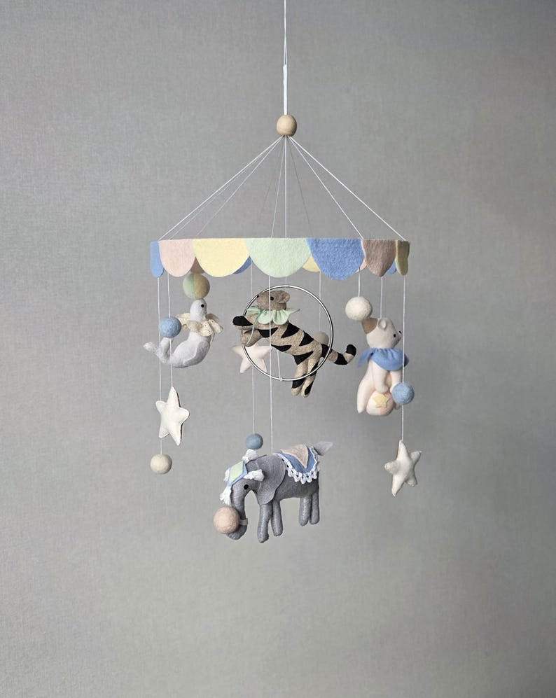 May include: A whimsical baby mobile featuring felt circus animals, including a tiger, elephant, seal, and bear. The mobile has a pastel-colored canopy and is suspended by strings with star and ball accents. The overall design is soft and inviting.