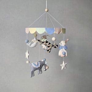 May include: A whimsical baby mobile featuring felt circus animals. The mobile includes a gray elephant, a tiger jumping through a hoop, a seal, and a bear. Pastel colors of blue, yellow, and pink are used throughout the design, with white stars and spheres.