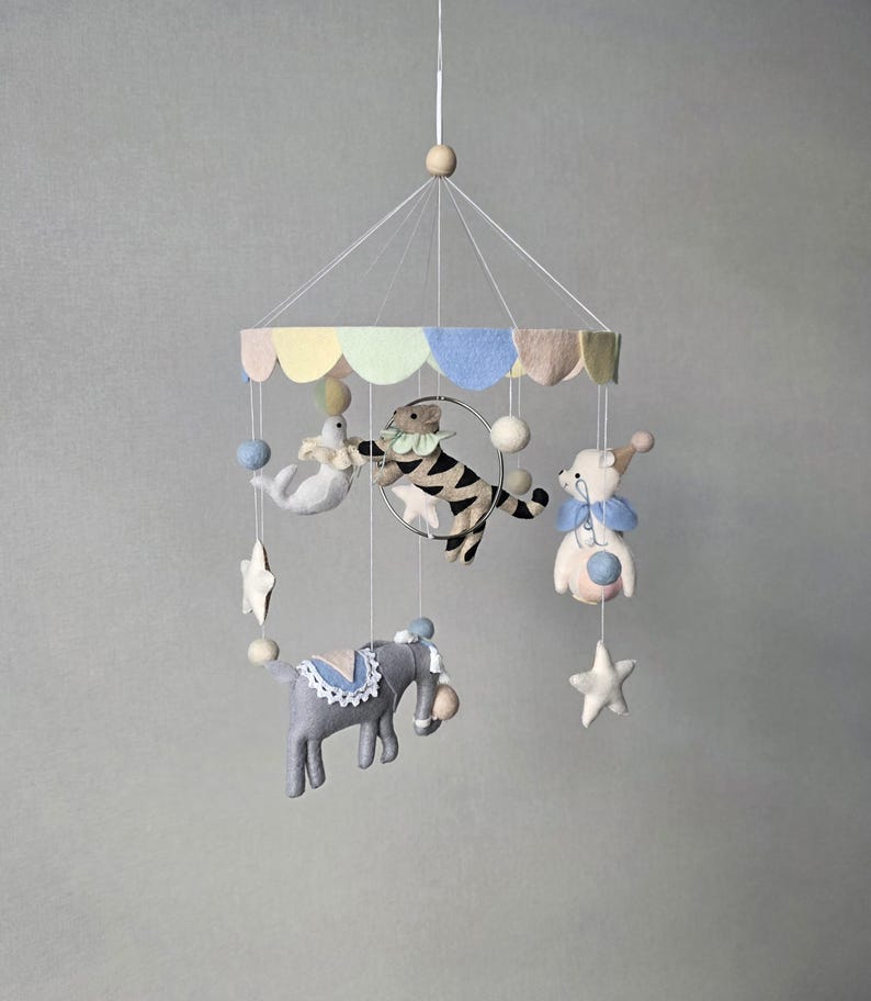 May include: A whimsical baby mobile featuring felt circus animals. The mobile includes a gray elephant, a tiger jumping through a hoop, a seal, and a bear. Pastel colors of blue, yellow, and pink are used throughout the design, with white stars and spheres.