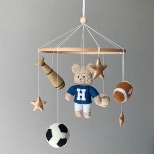 May include: A baby mobile featuring a wooden ring with hanging felt sports-themed toys. Includes a teddy bear in a blue shirt with a white "H", a baseball bat, stars, a soccer ball, a baseball, and a football. The toys are in shades of brown, white, and gold.