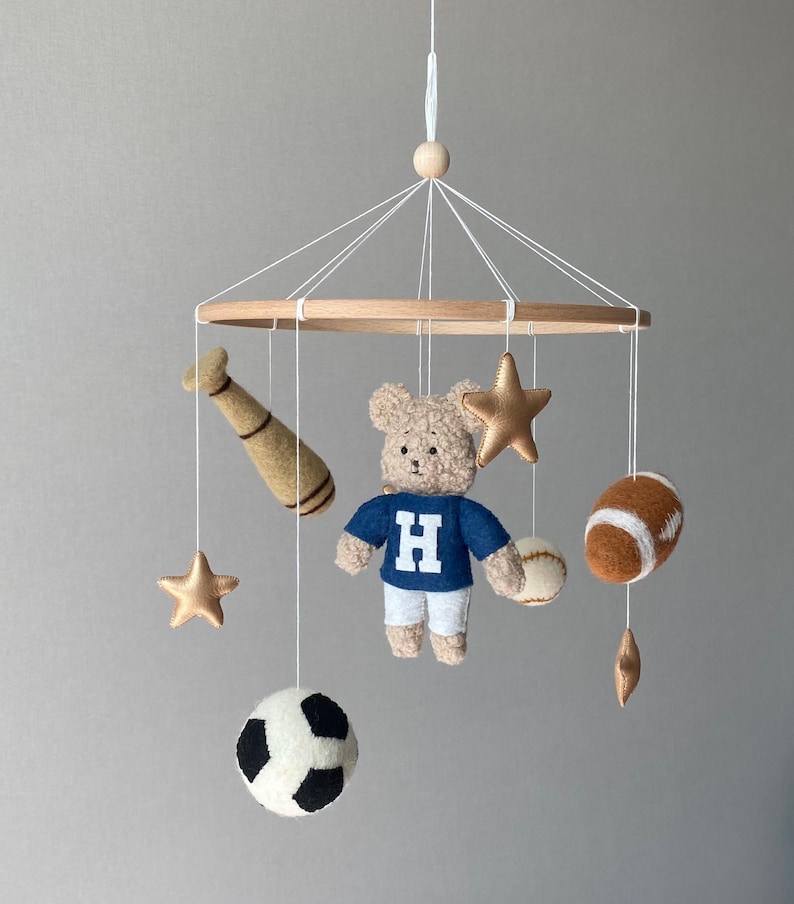 May include: A baby mobile featuring a wooden ring with hanging felt sports-themed toys. Includes a teddy bear in a blue shirt with a white "H", a baseball bat, stars, a soccer ball, a baseball, and a football. The toys are in shades of brown, white, and gold.