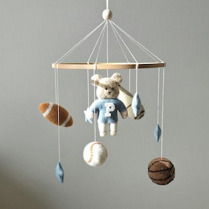 May include: A baby mobile featuring a wooden frame with hanging felt sports-themed toys. Includes a football, baseball, basketball, and a teddy bear wearing a blue sweater with a white "P" on it. The mobile is suspended by white strings.