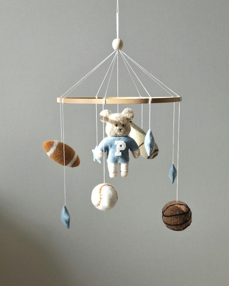 May include: A baby mobile featuring a wooden frame with hanging felt sports-themed toys. Includes a football, baseball, basketball, and a teddy bear wearing a blue sweater with a white "P" on it. The mobile is suspended by white strings.