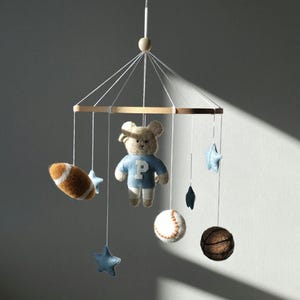 May include: A baby mobile featuring a teddy bear wearing a blue shirt with a white "P", a football, a baseball, a basketball, and blue stars. The mobile is suspended from a wooden frame with white strings.