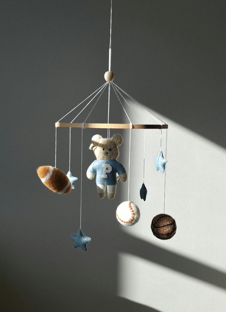 May include: A baby mobile featuring a teddy bear wearing a blue shirt with a white "P", a football, a baseball, a basketball, and blue stars. The mobile is suspended from a wooden frame with white strings.
