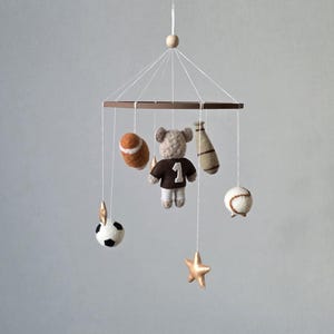 May include: A baby mobile featuring a brown wooden frame with hanging sports-themed felt ornaments. Includes a football, baseball, soccer ball, a bear wearing a jersey with the number 1, a baseball bat, and gold stars.