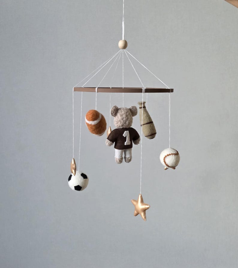 May include: A baby mobile featuring a brown wooden frame with hanging sports-themed felt ornaments. Includes a football, baseball, soccer ball, a bear wearing a jersey with the number 1, a baseball bat, and gold stars.