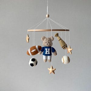 May include: A baby mobile featuring a teddy bear wearing a blue shirt with a white "H", a football, a soccer ball, a baseball, a baseball bat, and gold stars. The mobile is suspended from a wooden ring.