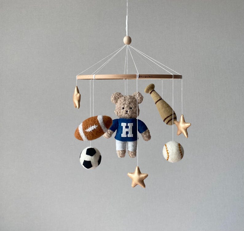 May include: A baby mobile featuring a teddy bear wearing a blue shirt with a white "H", a football, a soccer ball, a baseball, a baseball bat, and gold stars. The mobile is suspended from a wooden ring.