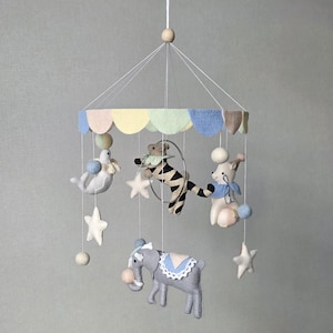 May include: A whimsical baby mobile featuring felt circus animals. The mobile includes a gray elephant, a bear, a seal, and a rabbit, all suspended from a colorful scalloped canopy. White stars and pom-poms add to the playful design.