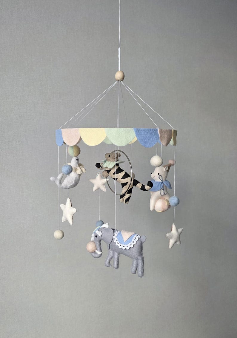 May include: A whimsical baby mobile featuring felt circus animals. The mobile includes a gray elephant, a bear, a seal, and a rabbit, all suspended from a colorful scalloped canopy. White stars and pom-poms add to the playful design.