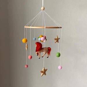 K&ouml;nnte beinhalten: Handgefertigtes Baby-Mobile mit Holzring und h&auml;ngenden Filzelementen. Das Mobile zeigt ein braunes Pferd mit roter M&auml;hne und Schweif, goldene Sterne, bunte Filzb&auml;lle und einen Ballon-Cluster. Aufgeh&auml;ngt an wei&szlig;en Schn&uuml;ren.