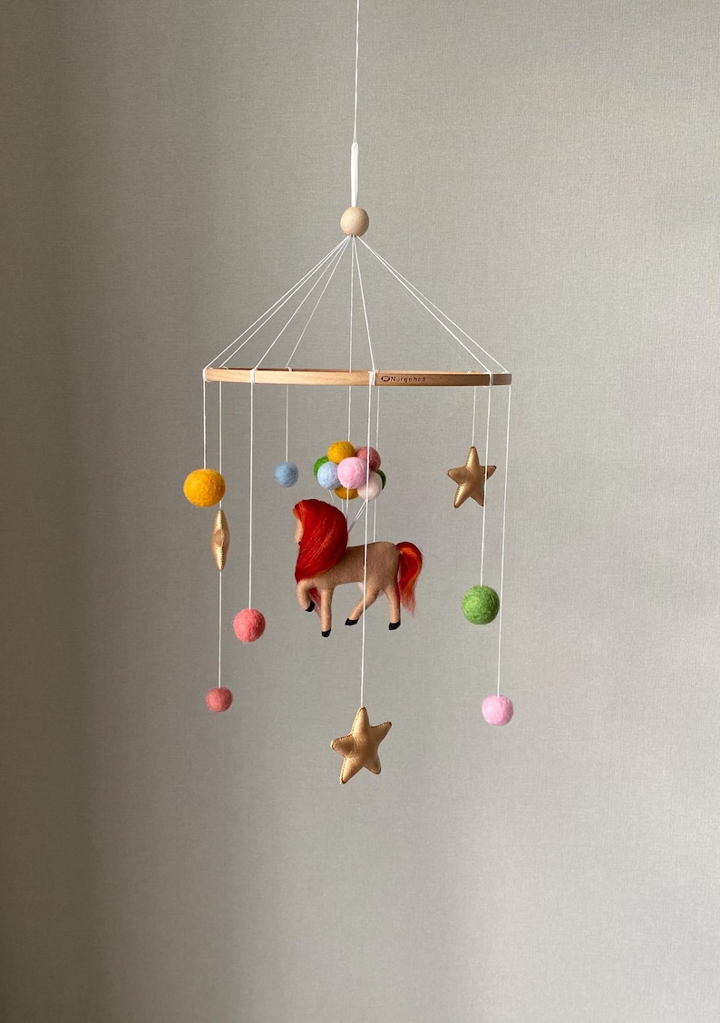 K&ouml;nnte beinhalten: Handgefertigtes Baby-Mobile mit Holzring und h&auml;ngenden Filzelementen. Das Mobile zeigt ein braunes Pferd mit roter M&auml;hne und Schweif, goldene Sterne, bunte Filzb&auml;lle und einen Ballon-Cluster. Aufgeh&auml;ngt an wei&szlig;en Schn&uuml;ren.