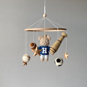 May include: A baby mobile featuring a teddy bear wearing a blue sweater with a white "H", a baseball bat, a football, a baseball, a soccer ball, and gold stars. The mobile is suspended from a wooden ring.