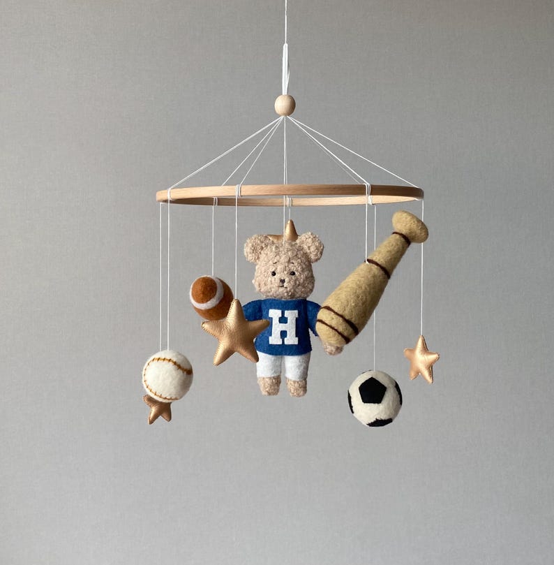 May include: A baby mobile featuring a teddy bear wearing a blue sweater with a white "H", a baseball bat, a football, a baseball, a soccer ball, and gold stars. The mobile is suspended from a wooden ring.