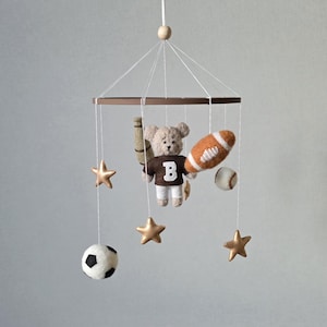 May include: A baby mobile featuring a teddy bear wearing a brown jersey with a white "B", holding a baseball bat and a football. The mobile also includes a baseball, soccer ball, and gold star ornaments. The mobile is suspended from a brown ring.
