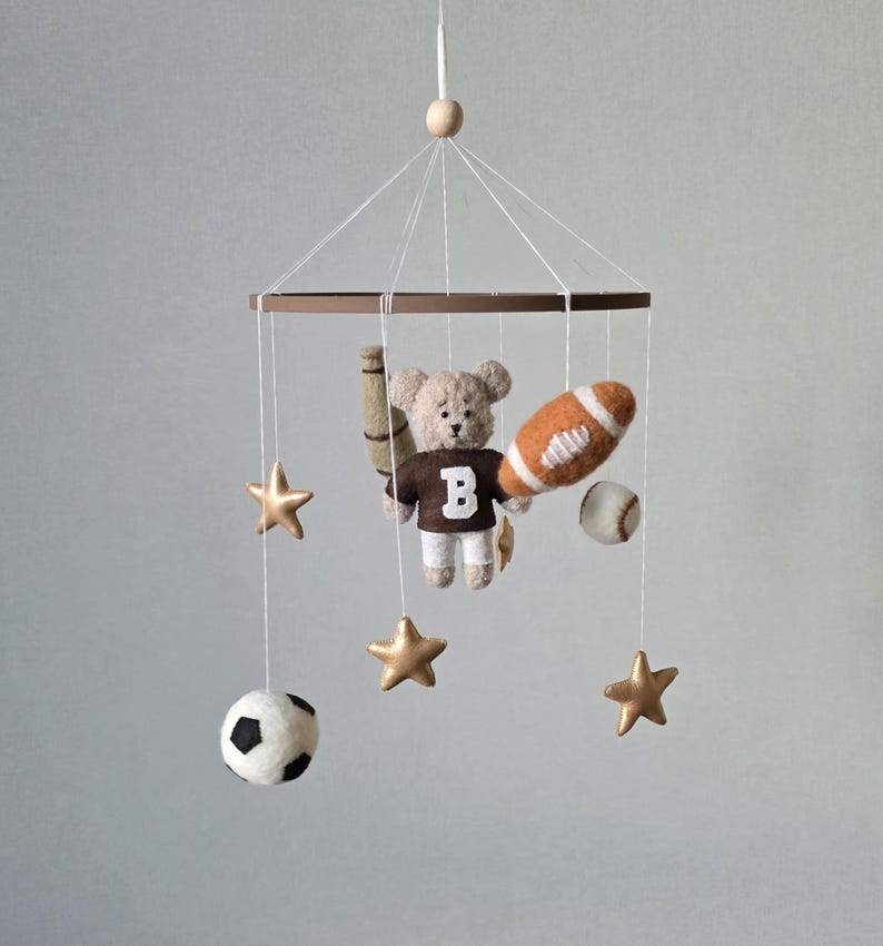 May include: A baby mobile featuring a teddy bear wearing a brown jersey with a white "B", holding a baseball bat and a football. The mobile also includes a baseball, soccer ball, and gold star ornaments. The mobile is suspended from a brown ring.