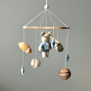 May include: A baby mobile featuring a wooden ring with hanging felt sports-themed toys. Includes a football, baseball, basketball, and a bear wearing a blue jersey with the number 1. Decorative blue star shapes are also included.