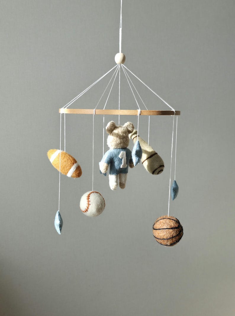 May include: A baby mobile featuring a wooden ring with hanging felt sports-themed toys. Includes a football, baseball, basketball, and a bear wearing a blue jersey with the number 1. Decorative blue star shapes are also included.