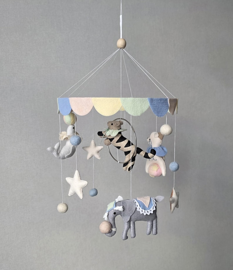 May include: A whimsical baby mobile featuring felt circus animals. The mobile includes a gray elephant, a zebra in a hoop, and a bear. Pastel colors are used throughout, with stars and pom-poms suspended from the top.