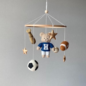 May include: A baby mobile featuring a wooden ring with hanging felt sports-themed ornaments. Includes a teddy bear wearing a blue jersey with a white "H", a baseball bat, a football, a soccer ball, a baseball, and gold stars. The mobile is suspended by white strings.