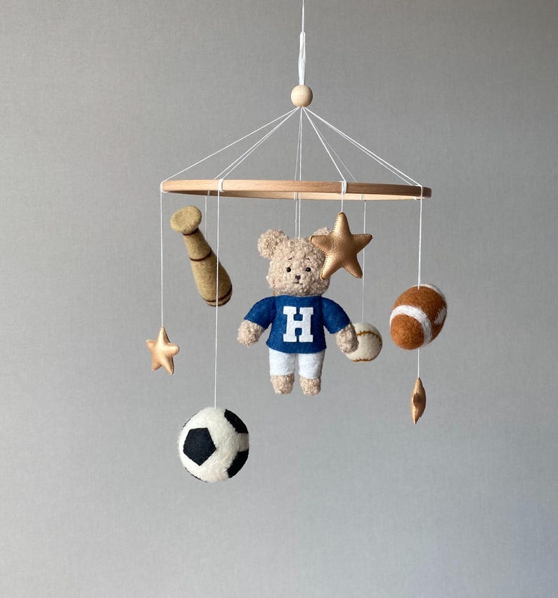 May include: A baby mobile featuring a wooden ring with hanging felt sports-themed ornaments. Includes a teddy bear wearing a blue jersey with a white "H", a baseball bat, a football, a soccer ball, a baseball, and gold stars. The mobile is suspended by white strings.