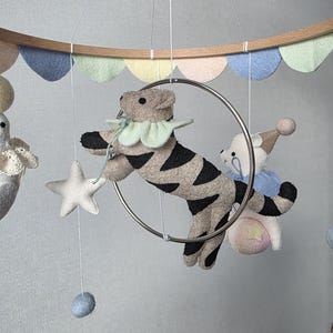 May include: A baby mobile featuring felt animal figures, including a tiger with black stripes, a bear, and a star. The mobile has a wooden ring with pastel-colored felt cutouts and is suspended by strings.