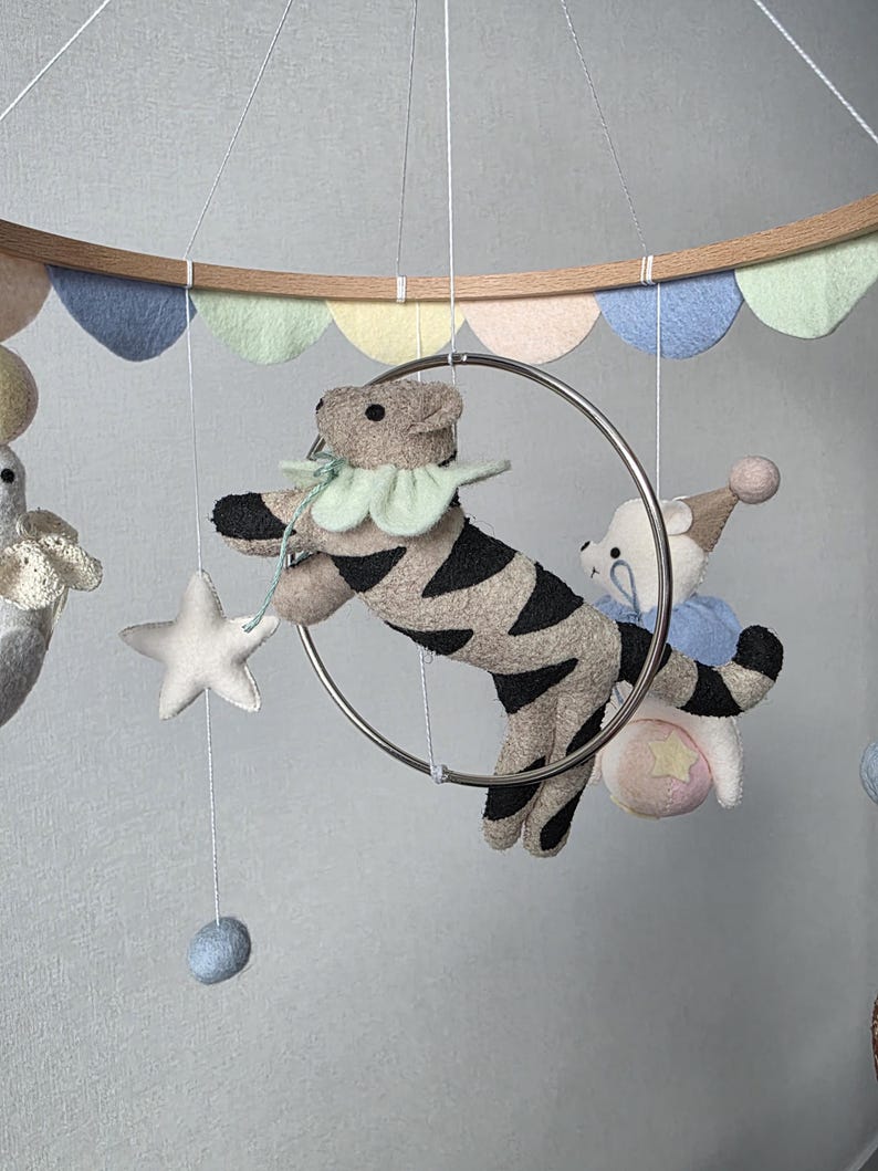 May include: A baby mobile featuring felt animal figures, including a tiger with black stripes, a bear, and a star. The mobile has a wooden ring with pastel-colored felt cutouts and is suspended by strings.