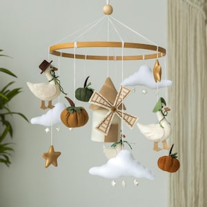 May include: A white and brown felt baby mobile with a windmill, two geese, clouds, stars, and pumpkins. The mobile is hanging from a wooden ring.