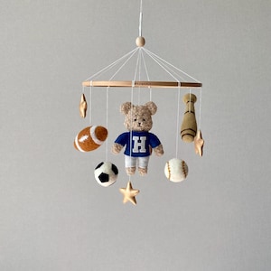 May include: A baby mobile featuring a teddy bear wearing a blue shirt with a white "H", surrounded by felt sports-themed ornaments. Includes a football, soccer ball, baseball, and a baseball bat, plus gold stars.