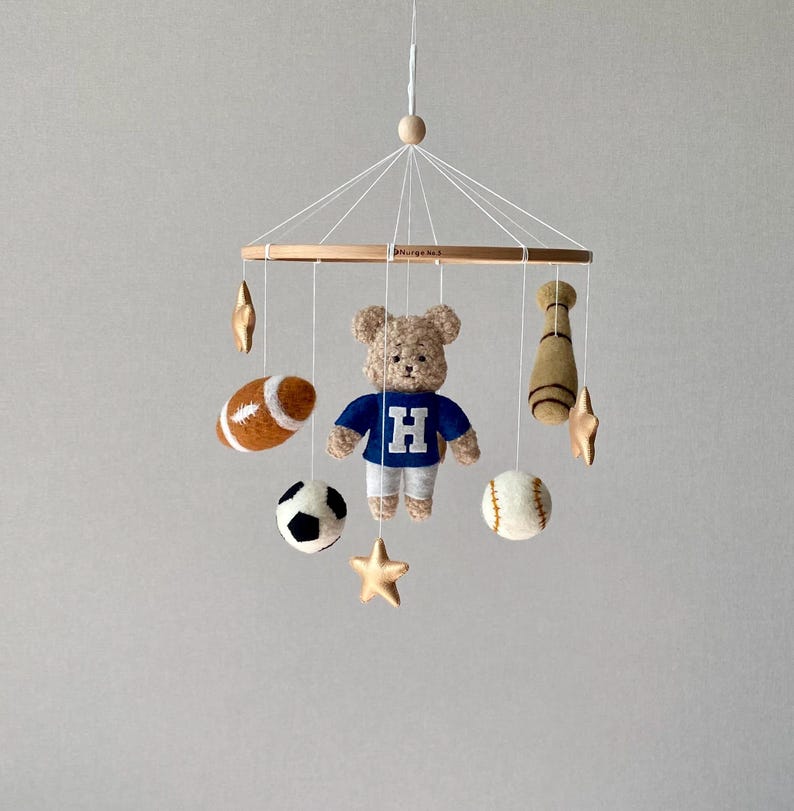 May include: A baby mobile featuring a teddy bear wearing a blue shirt with a white "H", surrounded by felt sports-themed ornaments. Includes a football, soccer ball, baseball, and a baseball bat, plus gold stars.