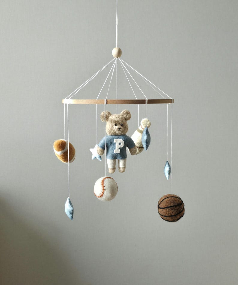 May include: A baby mobile featuring a teddy bear wearing a blue sweater with a white "P" and sports-themed felt ornaments. The mobile includes a baseball, basketball, football, and star-shaped and diamond-shaped accents, all suspended from a wooden ring.