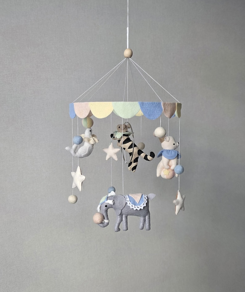 May include: A handmade baby mobile featuring felt circus animals, including an elephant, bear, zebra, and seal. The mobile has a pastel-colored canopy and star accents, suspended from a wooden bead.