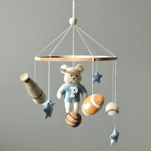 May include: A baby mobile featuring a teddy bear wearing a blue shirt with a white "P", a baseball bat, a football, a basketball, a baseball, and blue star shapes. The mobile is suspended from a wooden ring.