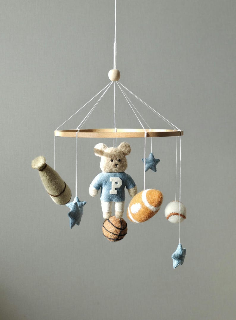May include: A baby mobile featuring a teddy bear wearing a blue shirt with a white "P", a baseball bat, a football, a basketball, a baseball, and blue star shapes. The mobile is suspended from a wooden ring.