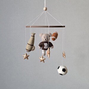 May include: A baby mobile featuring a teddy bear wearing a brown jersey with a white "B", a football, a soccer ball, a megaphone, and gold stars. The mobile is suspended from a wooden frame with a wooden bead at the top.
