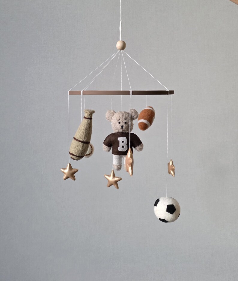 May include: A baby mobile featuring a teddy bear wearing a brown jersey with a white "B", a football, a soccer ball, a megaphone, and gold stars. The mobile is suspended from a wooden frame with a wooden bead at the top.
