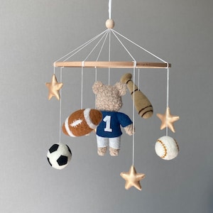 May include: A baby mobile featuring a teddy bear wearing a blue jersey with the number 1, a football, baseball, soccer ball, baseball bat, and gold stars. The mobile is suspended from a wooden frame.