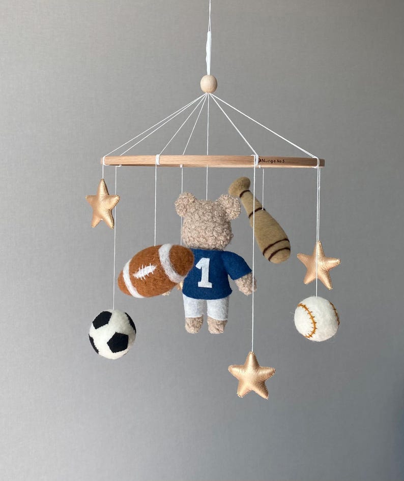 May include: A baby mobile featuring a teddy bear wearing a blue jersey with the number 1, a football, baseball, soccer ball, baseball bat, and gold stars. The mobile is suspended from a wooden frame.