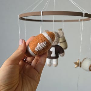 May include: A handmade baby mobile featuring a felt football, a teddy bear wearing a jersey with the number 1, a baseball, and gold stars. The mobile has a brown wooden ring and is suspended by white strings.