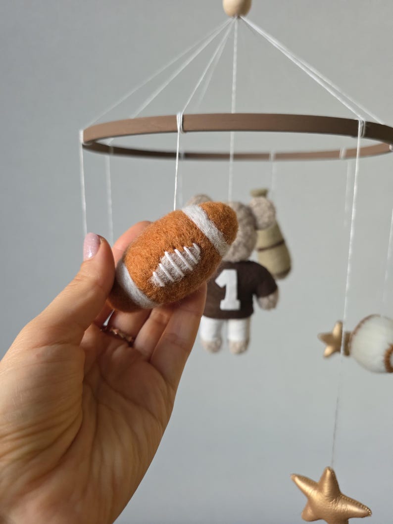 May include: A handmade baby mobile featuring a felt football, a teddy bear wearing a jersey with the number 1, a baseball, and gold stars. The mobile has a brown wooden ring and is suspended by white strings.
