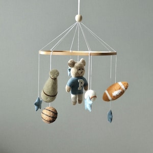 May include: A baby mobile featuring a wooden ring with hanging felt sports-themed toys. Includes a teddy bear in a blue shirt, a football, a baseball, a basketball, and star-shaped accents. The mobile is suspended by white strings.