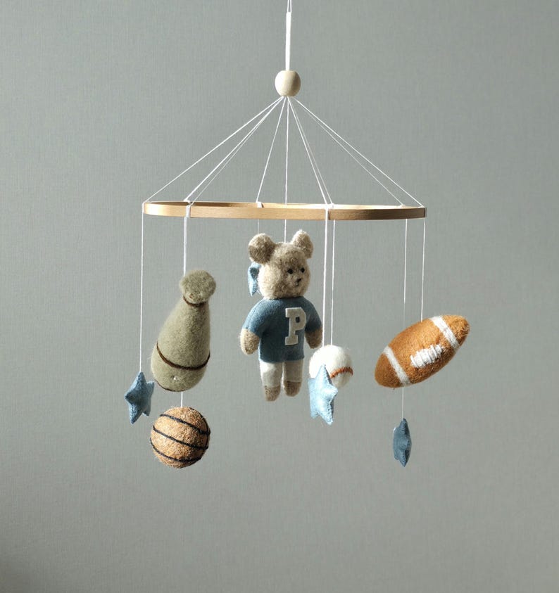May include: A baby mobile featuring a wooden ring with hanging felt sports-themed toys. Includes a teddy bear in a blue shirt, a football, a baseball, a basketball, and star-shaped accents. The mobile is suspended by white strings.