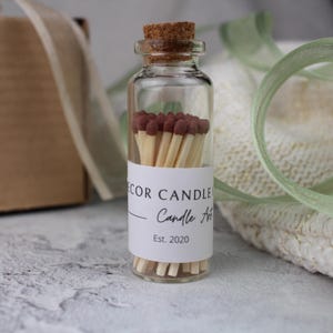 Peut inclure: Un petit bocal en verre avec un couvercle en li&egrave;ge rempli d'allumettes en bois avec des embouts bruns. Le bocal porte une &eacute;tiquette blanche sur laquelle est inscrit "Decor Candle Co. Candle Art Est. 2020".