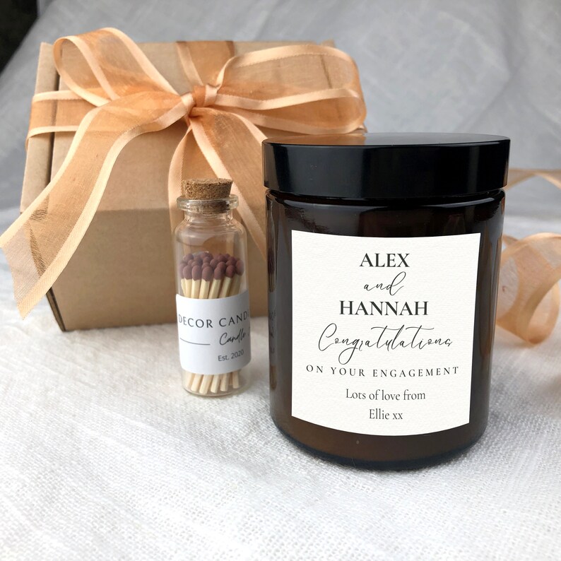 May include: A brown glass candle jar with a black lid and a white label that reads "ALEX and HANNAH Congratulations ON YOUR ENGAGEMENT Lots of love from Ellie xx". The candle is packaged in a gift box with a brown ribbon and a small glass jar of matches.