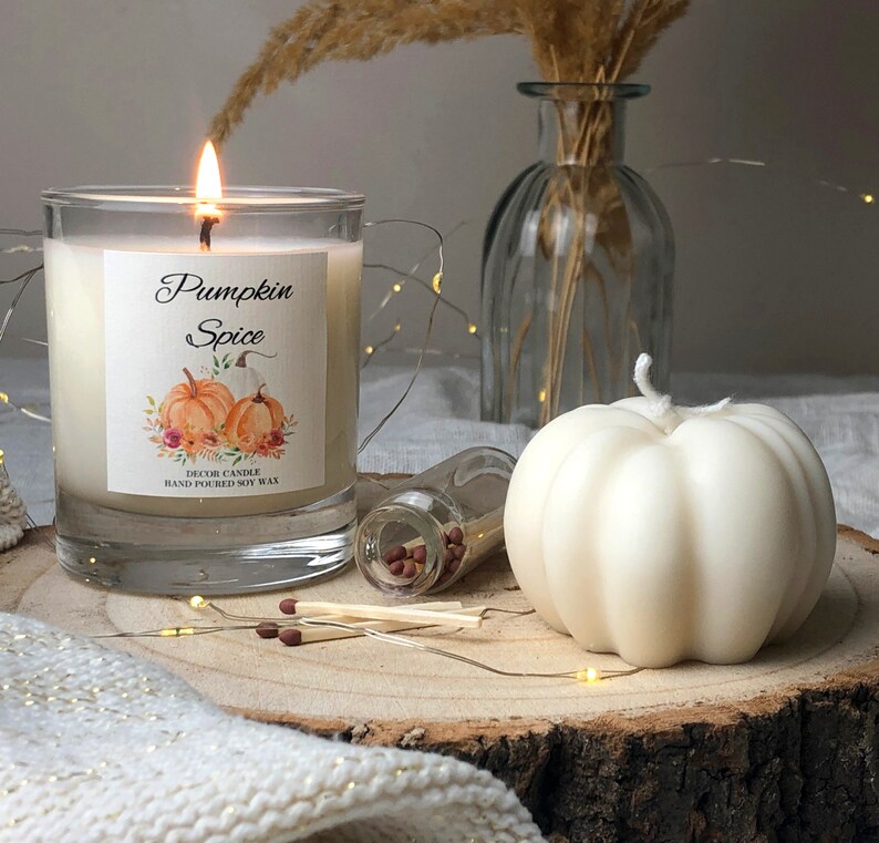 May include: A white pumpkin-shaped candle with a label that reads "Pumpkin Spice" and "Decor Candle Hand Poured Soy Wax". The candle is sitting on a wooden surface with a glass jar of matches and a clear glass vase with dried grass.