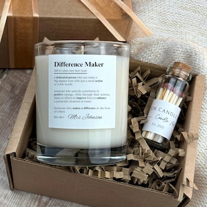 May include: A white candle in a glass jar with a brown paper label that reads "Difference Maker" and a definition of the word. The candle is in a brown box with shredded paper. A small glass bottle with matches is next to the candle. The bottle has a label that reads "For Candle - Candle Art - Est. 2020".