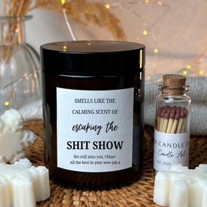 Peut inclure: Un bocal de bougie noir avec une &eacute;tiquette blanche qui indique "Smells like the calming scent of escaping the shit show. We will miss you, Chloe! All the best in your new job x". La bougie est pos&eacute;e sur une surface en osier tress&eacute;. Il y a un petit bocal en verre avec des allumettes &agrave; c&ocirc;t&eacute; de la bougie.